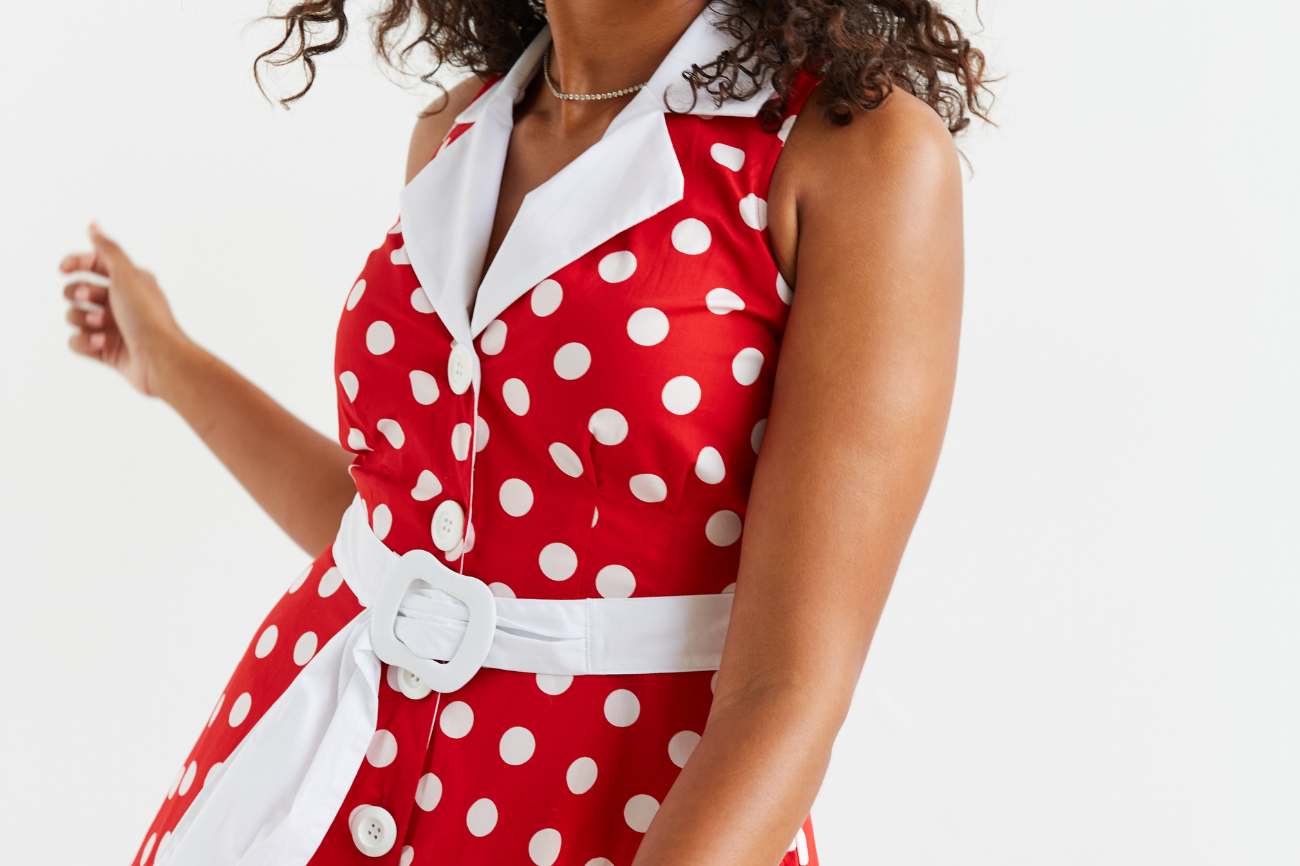 Women standing in front of a white wall , she is facing on a slight angle and looking down over her shoulder towards her outstretched hand.  Her dark curly hair is loosely flowing over her shoulders. She is wearing a red and white polka dot midi dress, the image is cropped and you can see 3/4 of the dress.  She has a wry smile.
