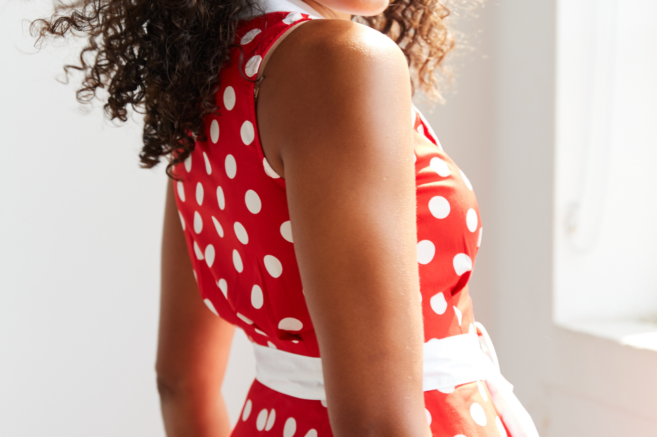 Women standing beside a window , she is facing sideways and looking over shoulder with dark curly hair blowing. She is wearing a red and white polka dot midi dress