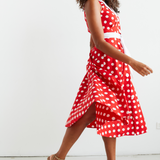 Women standing in front of a white wall , she is facing on a slight angle and looking directly over her shoulder towards the viewer.  Her dark curly hair is loosely flowing over her shoulders. She is wearing a red and white polka dot midi dress, which she has twirled in and we can see the voluminous skirt of the dress