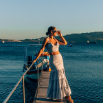  Crop Top and Maxi Skirt set in Blue and White Stripe 100% cotton seersucker deadstock fabric  worn by woman with long brown hair in a bun on a long pier surrounded by blue water, mountains and boats.  She has one hand up to her face touching her sunglasses and looking out towards the sun setting.