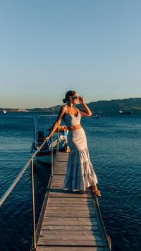  Crop Top and Maxi Skirt set in Blue and White Stripe 100% cotton seersucker deadstock fabric  worn by woman with long brown hair in a bun on a long pier surrounded by blue water, mountains and boats.  She has one hand up to her face touching her sunglasses and looking out towards the sun setting.